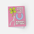 Pink card with a four-leaf clover, blue horseshoe, and white wishbone on a white background