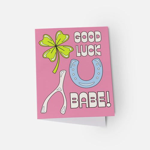 Pink card with a four-leaf clover, blue horseshoe, and white wishbone on a white background